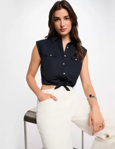 Top feminino azul escuro sem mangas com botões e bolso, calças brancas, mulher sentada