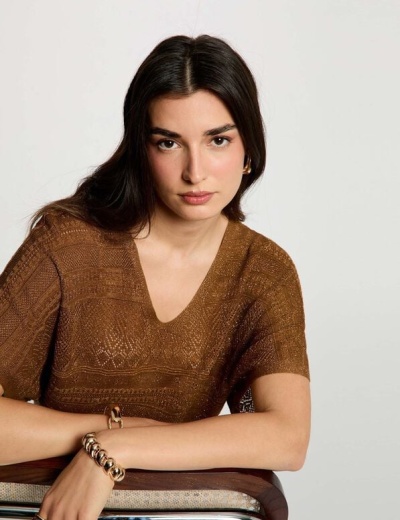 Mulher com blusa castanha de tricô e acessórios dourados sentada