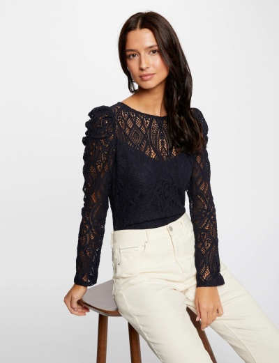 Modelo com blusa preta de malha rendada e calças claras sentada num banco de madeira