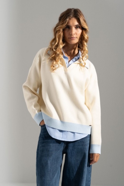 Mulher com camisola creme e azul claro sobre camisa azul, calças de ganga azuis, fundo branco.