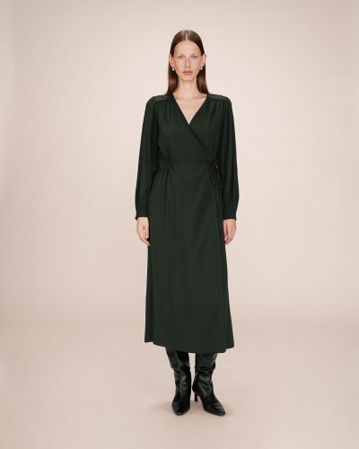 Vestido longo verde escuro de manga comprida com cinto, usado com botas pretas e fundo bege.