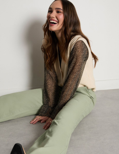 Mulher sentada vestindo blusa preta transparente com padrão de bolinhas brancas, colete creme de malha e calças verdes claras.