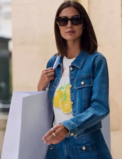 Mulher com casaco e calças de ganga azuis, t-shirt branca com limões, óculos de sol escuros e sacola branca