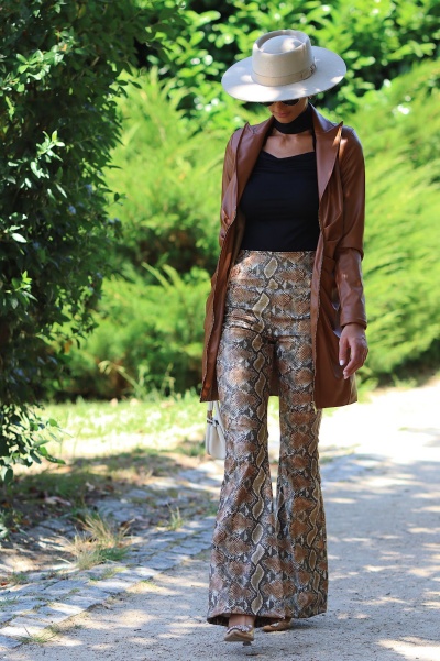Mulher com casaco castanho, calças de padrão pele de cobra e chapéu bege a caminhar numa zona exterior com vegetação.
