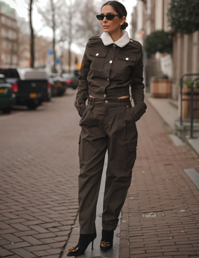 Conjunto castanho feminino com gola de pêlo branca, calças cargo, óculos de sol pretos e botas de salto alto.