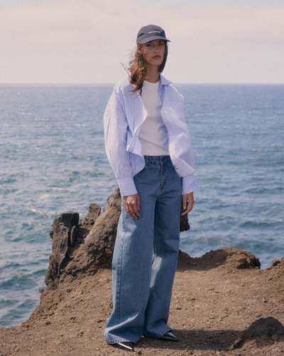 Mulher vestindo camisa azul de riscas, camisola branca, jeans largos azuis e boné azul escuro com texto, em pé perto do mar e rochas.