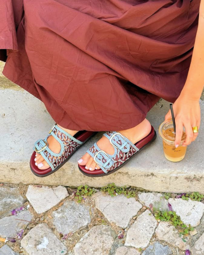 Sandálias femininas trançadas azul/castanho com fivelas prateadas e vestido castanho sentada em degrau com copo de bebida.