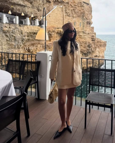 Mulher com roupa bege, chapéu estampado e bolsa bege numa varanda com vista para o mar