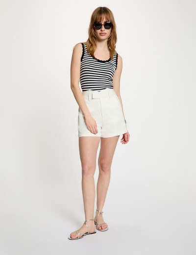 Mulher de pé com top às riscas preto e branco, calções brancos, sandálias prateadas e óculos de sol pretos sobre fundo branco.