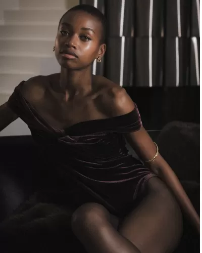 Mulher com body de veludo roxo escuro e joias douradas sentada em ambiente interior