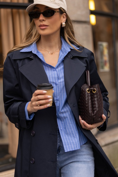 Mulher com trench coat azul, camisa riscada azul, jeans, óculos de sol pretos, boné bege e bolsa castanha de couro trançado segurando café.