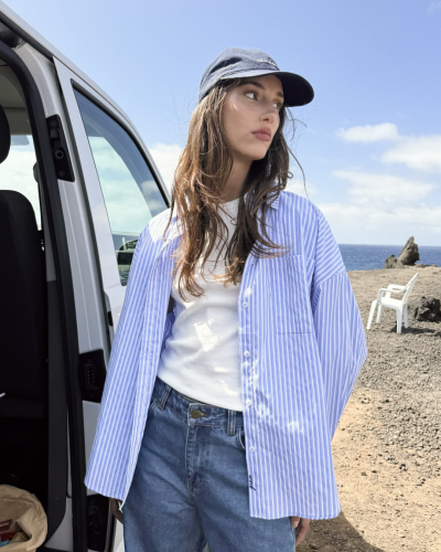 Mulher com camisa azul às riscas aberta, t-shirt branca, jeans e boné azul junto a carrinha numa paisagem costeira