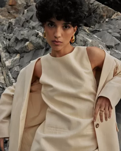 Modelo de mulher com vestido e blazer bege com riscas finas de branco, em ambiente rochoso.