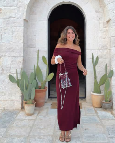 Mulher com vestido longo bordô e mala transparente com franjas em frente a entrada de pedra com cactos