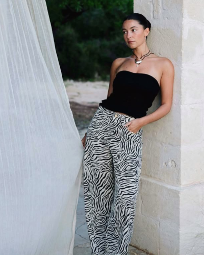 Mulher com top preto e calças de padrão zebra encostada em parede de pedra clara com cortina branca ao lado