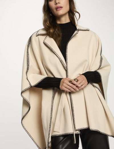 Capa bege com costuras pretas em ziguezague, camisola preta e calças de pele.