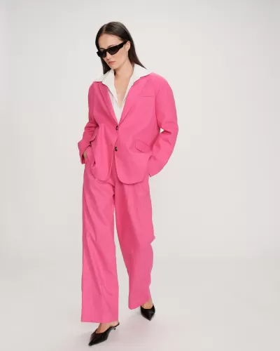 Mulher vestindo blazer e calças cor-de-rosa, camisa branca, sapatos pretos, óculos escuros, fundo branco