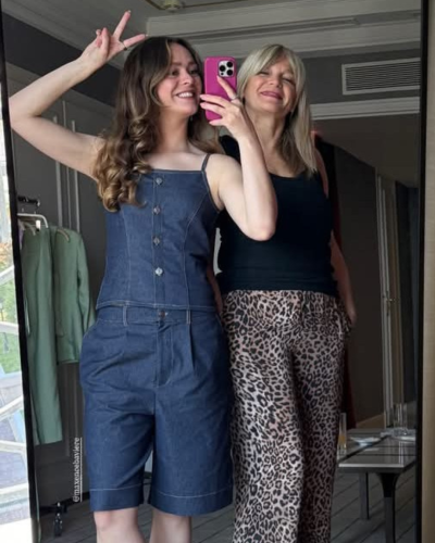 Duas mulheres tirando selfie no espelho com roupa casual, incluindo jardineira de ganga e calças de estampa animal.