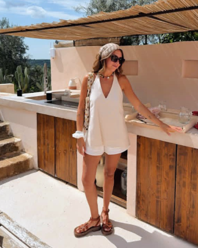Mulher com roupa de verão branca e sandálias castanhas numa cozinha exterior com armários de madeira.