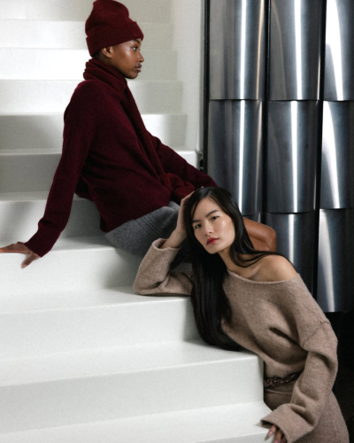 Duas mulheres com roupas de malha grosso bordô e castanho claro sentadas numa escada branca, fundo com paredes metálicas.