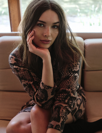 Mulher com vestido animal print castanho e preto sentada num sofá bege.