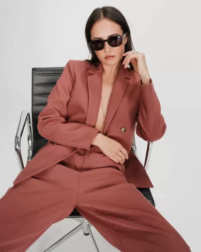 Mulher com fato castanho avermelhado e óculos sentada em cadeira de escritório preta.