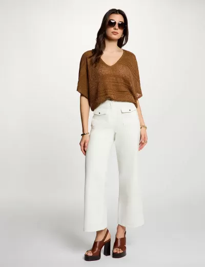 Mulher de blusa castanha, calças brancas e sapatos castanhos de salto em fundo branco