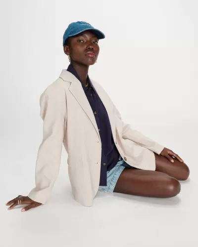 Mulher sentada com blazer bege, camisa azul escura, calções de ganga e boné azul com texto GRACE BLUE