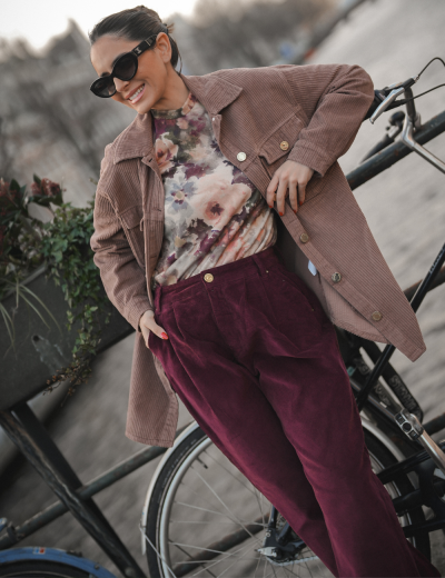 Mulher com jaqueta castanha, camisola floral e calças roxas apoiada numa bicicleta preta.