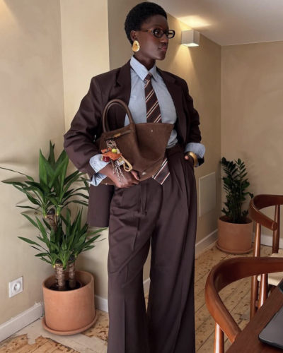 Mulher com blazer e calças castanho escuro, camisa azul, gravata riscada, mala castanha, plantas em vasos e cadeira de madeira ao lado.