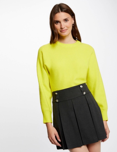 Mulher com camisola amarela e saia preta plissada em fundo branco