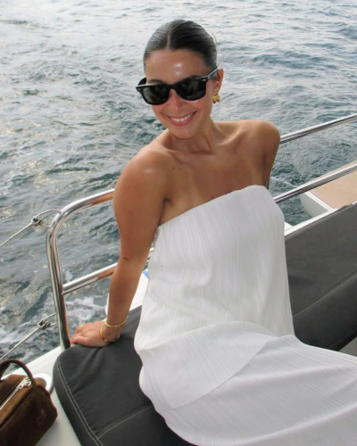 Mulher de vestido branco alças, óculos escuros e acessórios dourados sentado em banco de barco com mar ao fundo