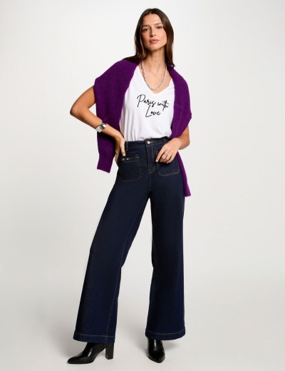 Mulher com calças largas azul escuras, t-shirt branca com texto, cardigan roxo e botas pretas em fundo branco