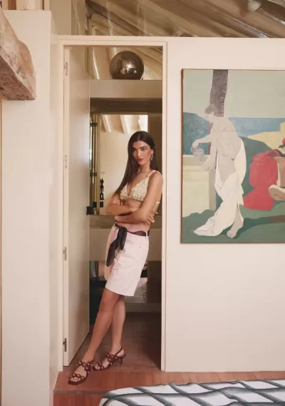 Mulher com top branco de renda e saia branca, com lenço na cintura, em sala com parede branca e pintura colorida