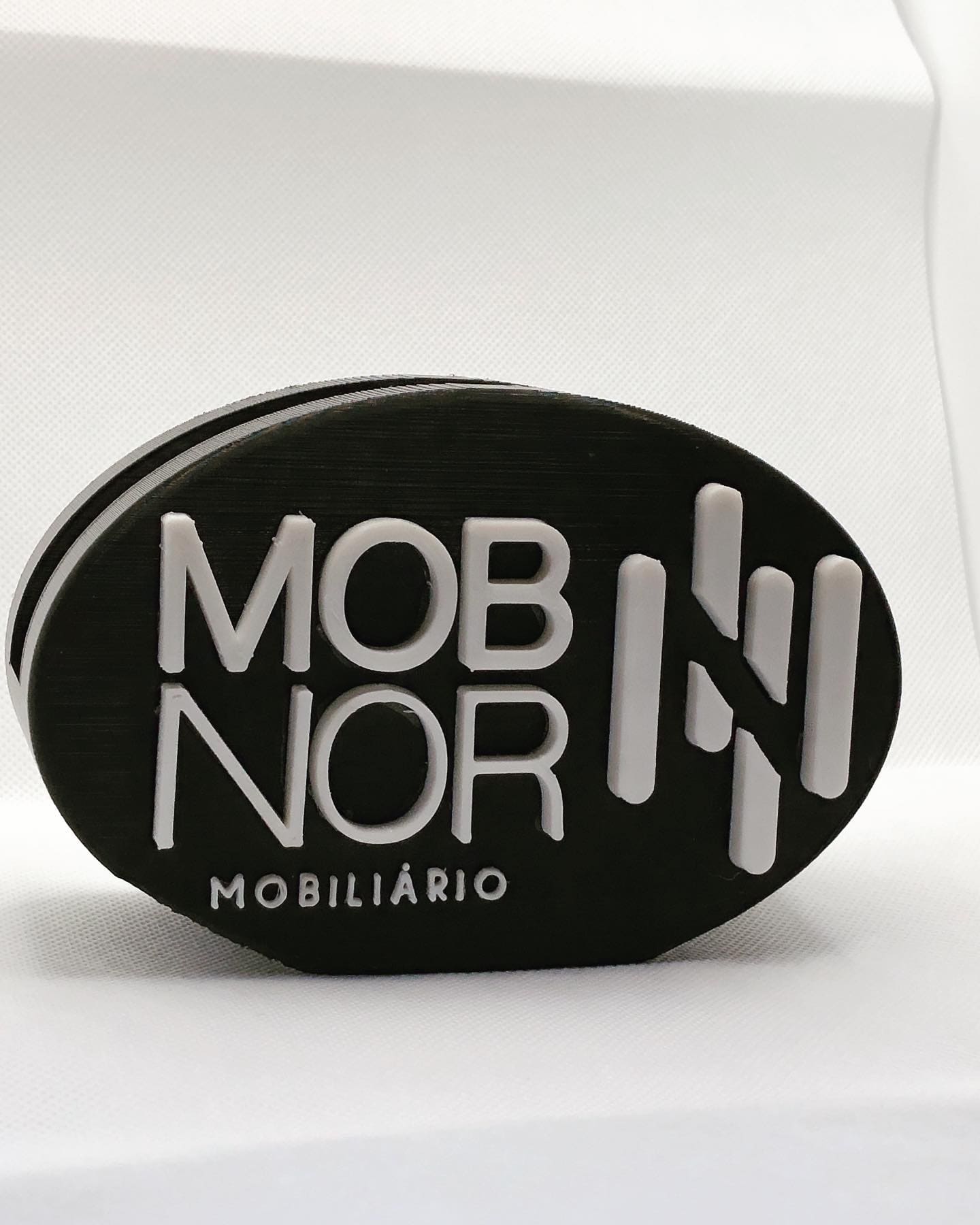 Serviço de Impressão 3D Personalizada – Transforme seus Modelos Digitais em Realidade Placa oval preta com texto branco MOB NOR MOBILIÁRIO sobre fundo branco
