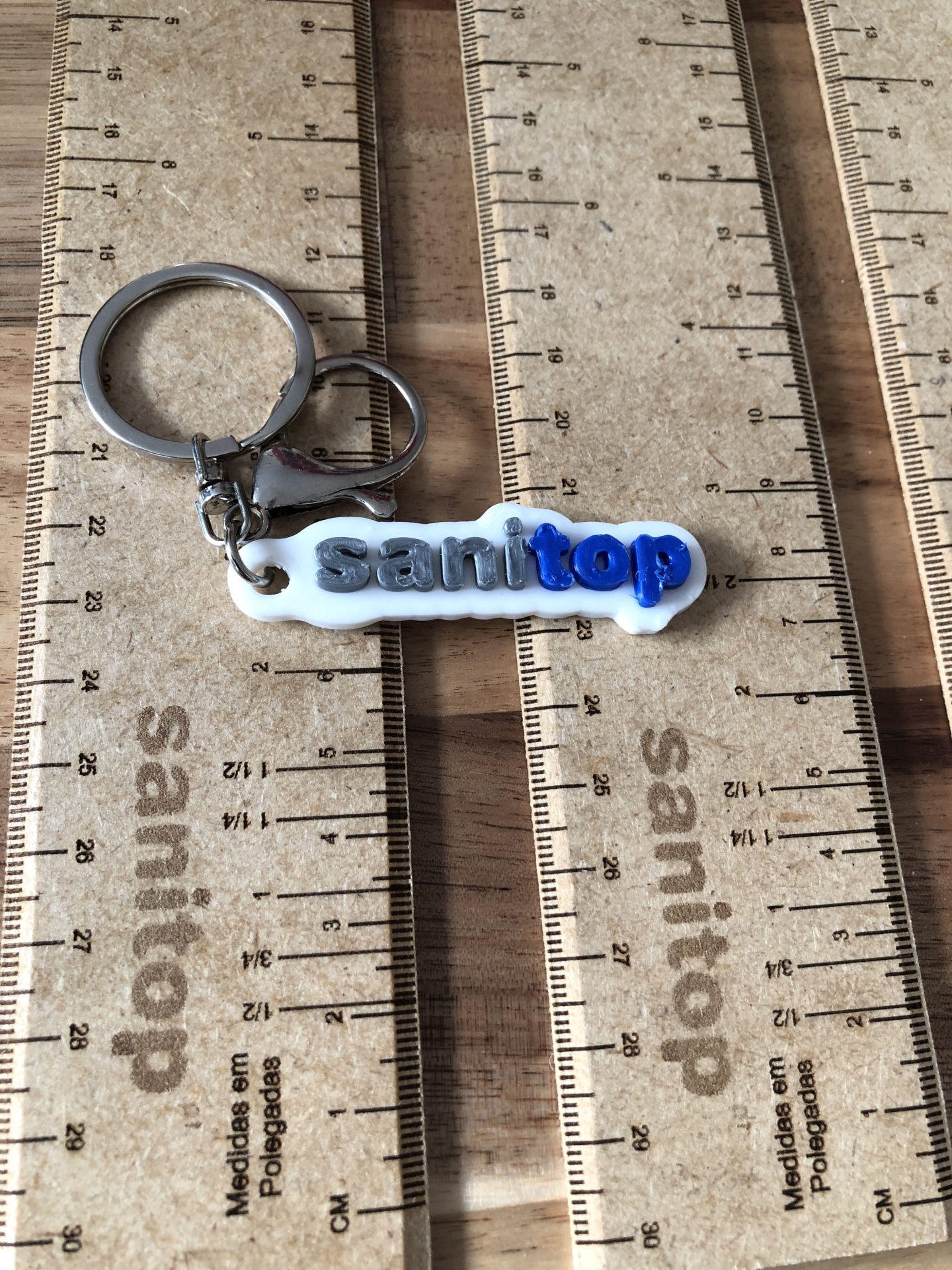Serviço de Impressão 3D Personalizada – Transforme seus Modelos Digitais em Realidade Chaveiro plastico branco com texto sanitop azul e preto sobre réguas de madeira.