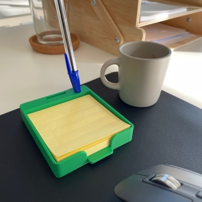 Bloco amarelo com suporte verde, caneta azul, chávena de café branca e rato de computador cinzento numa secretária branca
