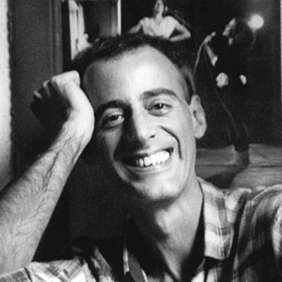 Homem sorridente com camisa xadrez e quadros artísticos ao fundo