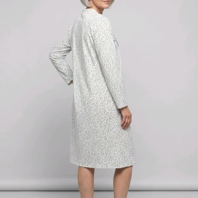 Mulher de cabelos grisalhos com vestido comprido de mangas compridas branco com padrões, chinelos brancos, fundo cinzento claro.