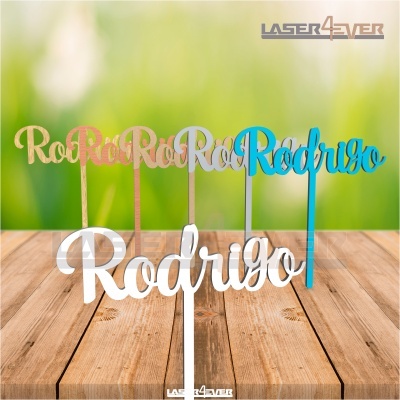 Cinco toppers de bolo com o nome 'Rodrigo' em diferentes cores, em cima de uma mesa de madeira, fundo desfocado verde.