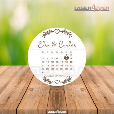 Calendário redondo com nomes, mês e dia destacado em coração castanho sobre bancada de madeira.