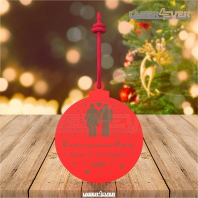 Ornamento de Natal vermelho personalizado com texto e figuras de casal, pendurado sobre madeira, com árvore de Natal ao fundo