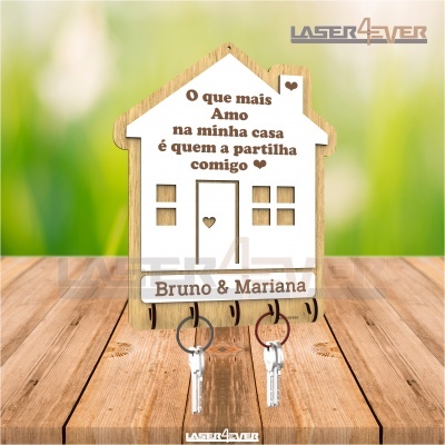 Porta-chaves em forma de casa com texto e nomes gravados, feito em madeira clara e branca, com duas chaves penduradas e fundo verde desfocado.
