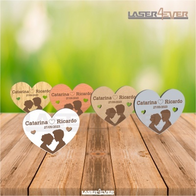 Seis corações de madeira com gravações de nomes, data e silhuetas, sobre mesa de madeira e fundo verde desfocado