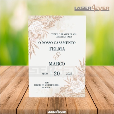 Convite de casamento branco com desenhos florais castanhos, texto em preto e castanho com data e nomes, sobre mesa de madeira.