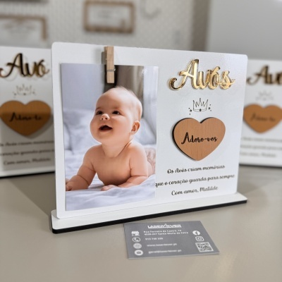 Porta-fotos branco e preto com palavra dourada Avós, coração de madeira e texto em português, com uma foto de bebé e cartões de visita ao fundo.