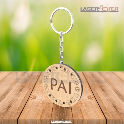 Chaveiro redondo de madeira com palavra 'PAI' e nomes gravados, pendurado sobre superfície de madeira clara