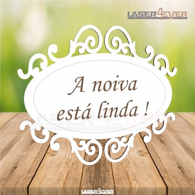 Placa decorativa branca oval com texto 'A noiva está linda !' em fundo verde desfocado