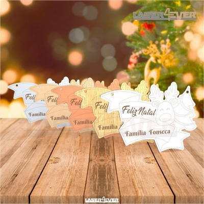 Etiquetas de Natal em madeira com formas de rena e árvore e texto gravado, sobre mesa de madeira com fundo natalício desfocado.