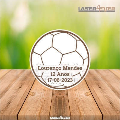 Objeto branco redondo com desenho de bola de futebol e texto comemorativo sobre madeira clara